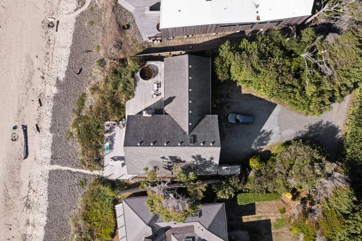 An exterior view of the property featuring the house nestled among lush greenery with a gravel driveway leading up. The beach is visible beyond, hinting at the close proximity to sand and surf, and the serene setting is ideal for exploring nature with pets.