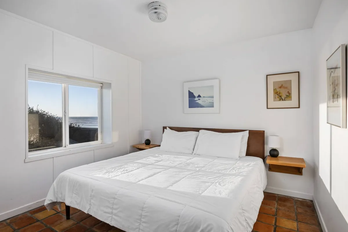 A tranquil bedroom with a plush bed, crisp white linens, a large window bringing in natural light, and calming wall art. The window offers a serene view, creating an inviting and restful atmosphere for relaxation or reading.