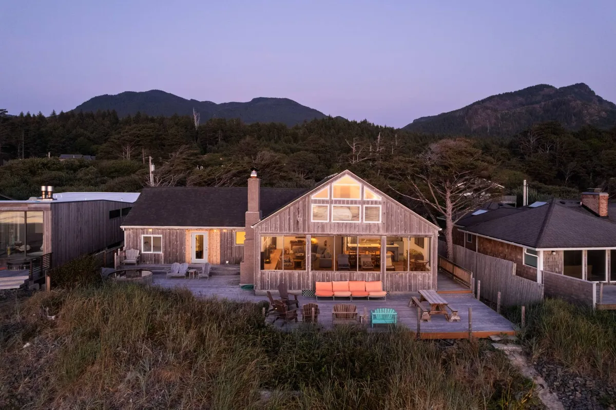The exterior of Casa Pacifica features large windows gleaming in the sunset, a spacious wooden deck with seating, and verdant mountain and tree views creating a tranquil backdrop. The inviting deck suggests ample space for pets and people to relax outdoors together.
