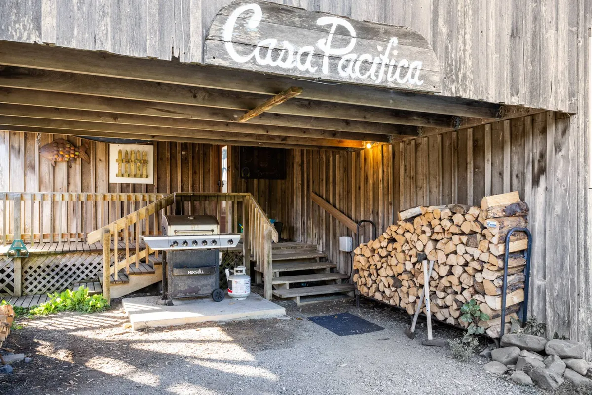 A rustic scene showing the wooden sign for 'Casa Pacifica,' an outdoor grill, stacked firewood, and a staircase leading toward the house. The setup suggests a welcoming entry point and a communal gathering spot after a day of exploration.