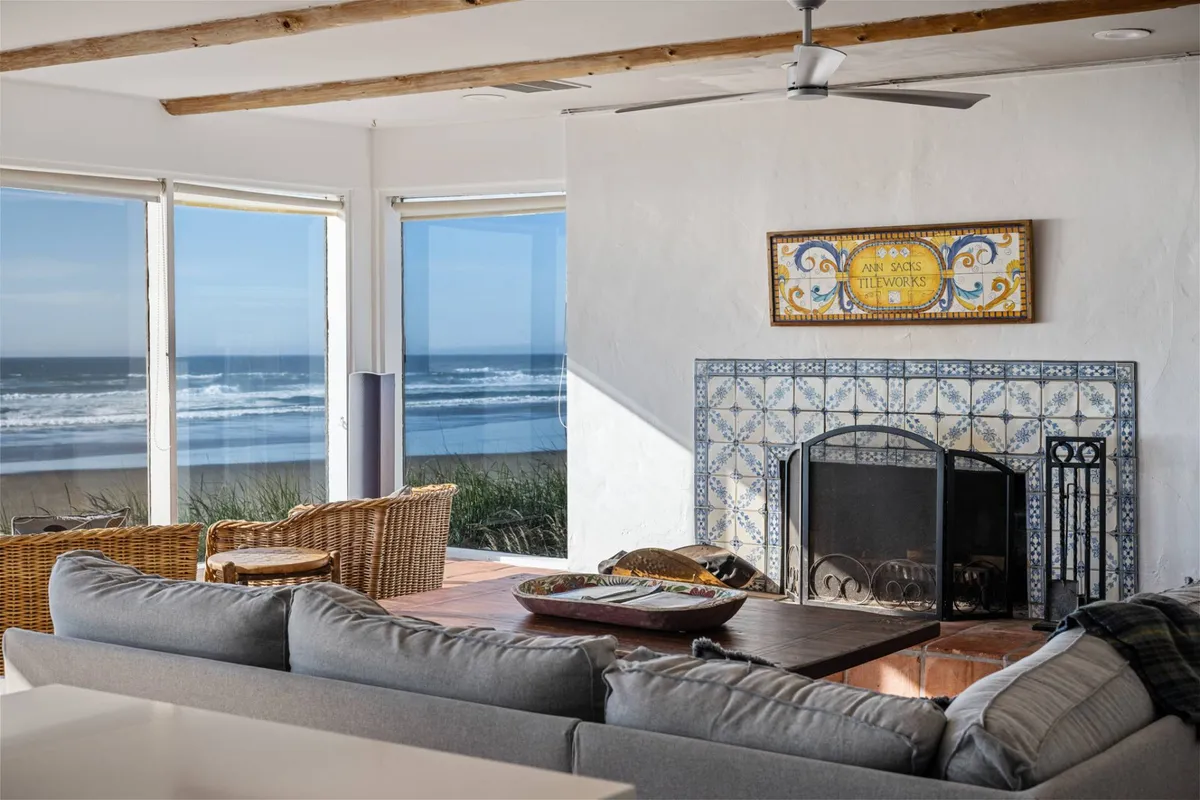 This image offers a close-up perspective of Casa Pacifica's inviting living area, with a plush sofa and woven chairs arranged around a tile-adorned wood-burning fireplace. Floor-to-ceiling windows frame the spectacular ocean views, and the setting radiates cozy comfort—perfect for relaxing, socializing, and enjoying storm-watching or sunny days.
