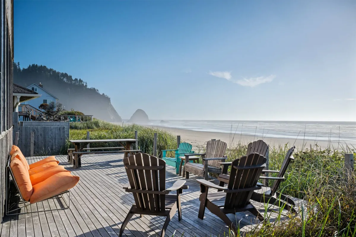 This image captures a deck with comfortable chairs positioned to overlook the ocean and beach, with the house visible nearby and an inviting orange sofa adding a pop of color. The expansive view of the sandy shoreline and rolling surf is front and center, suggesting easy access from the home to the beach for walks or play.