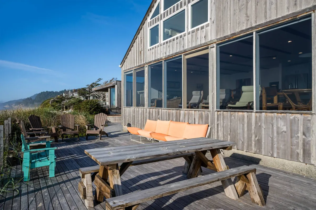A wide view of the home's expansive deck furnished with wooden outdoor tables and chairs, set against a backdrop of large glass windows and panoramic ocean views. The scene suggests space for group dining and socializing, with the ocean visible just beyond the deck.
