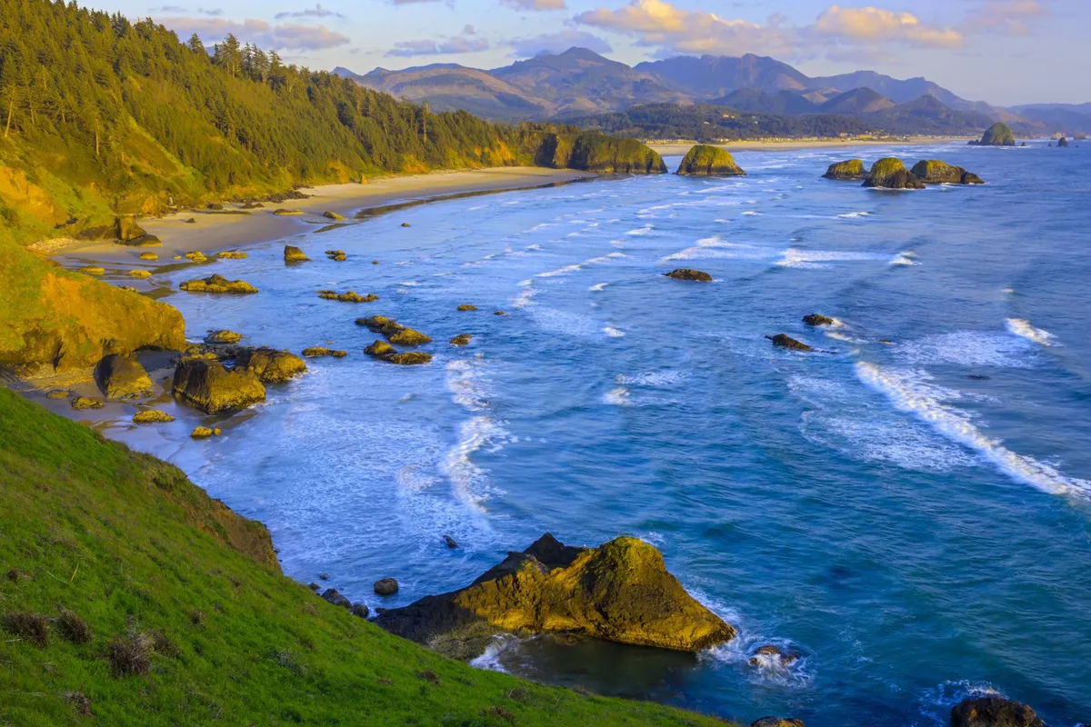 A dramatic coastal view featuring a rocky shoreline, waves crashing against the rocks, distant mountains, and lush green hills. The scene is wild and untamed, evoking the spirit of adventure and natural exploration. The area is ideal for hiking, wildlife spotting, and photography.