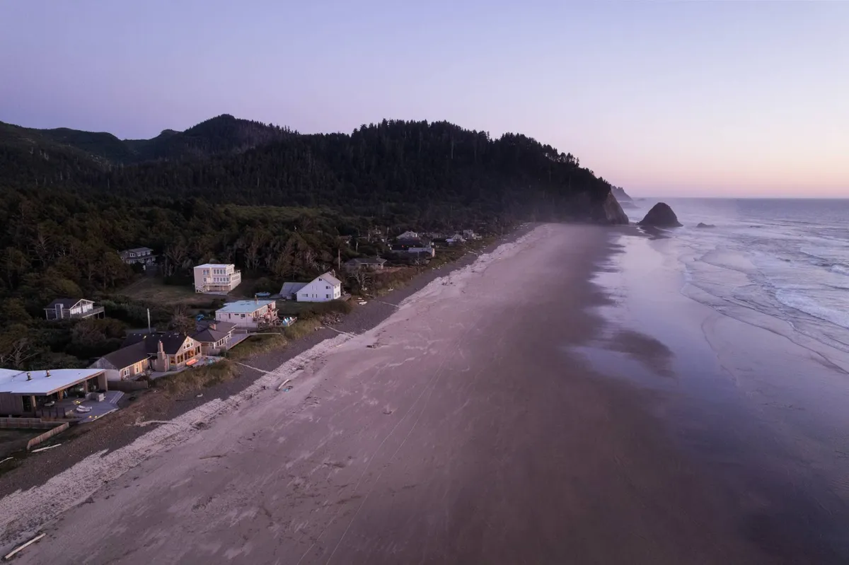 A panoramic view that includes a wide sandy beach, a calm ocean, a line of houses, and distant mountains. The scene is peaceful and expansive, suggesting both accessibility for visitors and the vastness of the region’s natural beauty.