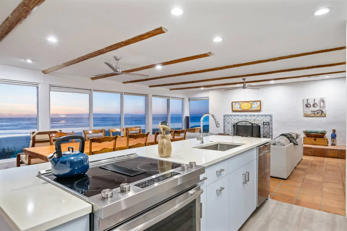 This image showcases the heart of Casa Pacifica's open-concept space, featuring a spacious kitchen island with a stovetop, large windows framing breathtaking ocean views, a generous dining table, and a cozy fireplace. The room seamlessly blends modern updates with warm, Spanish-inspired décor and is filled with natural light, inviting guests to cook, dine, and gather together.