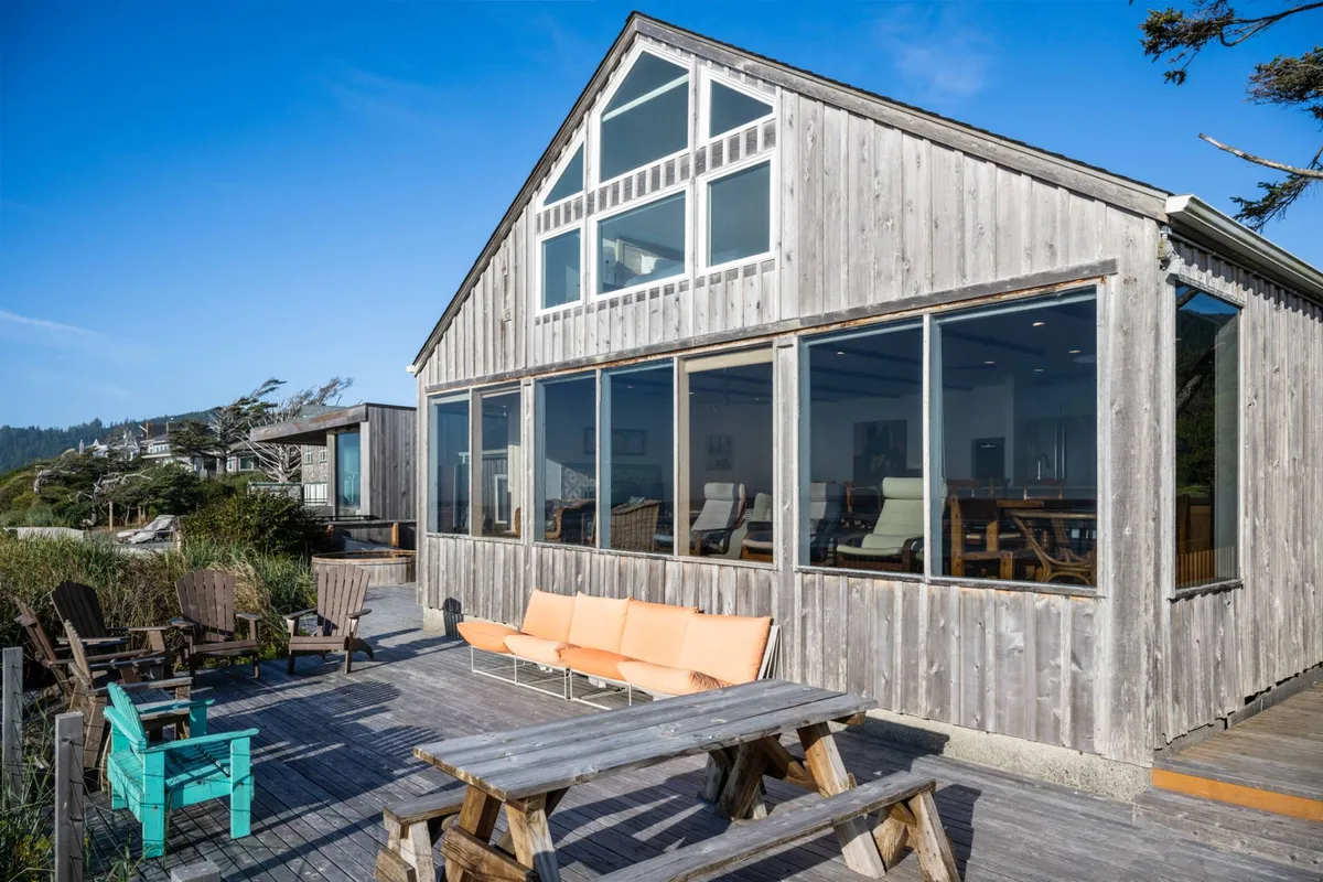 This image shows the exterior of Casa Pacifica, featuring large glass windows and a spacious wooden deck with outdoor seating. The scene is set against a coastal backdrop, inviting guests to relax in the sun or enjoy meals outdoors while taking in panoramic ocean views.