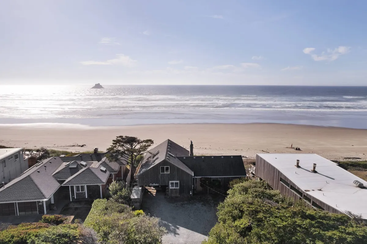 A sweeping oceanfront view encompassing the sandy beach, gentle waves, distant rock formations, and a row of charming houses facing the shore. The relaxed scene includes people beachcombing, sunbathing, and walking along the water's edge, with the vibrant sky hinting at a perfect sunset.