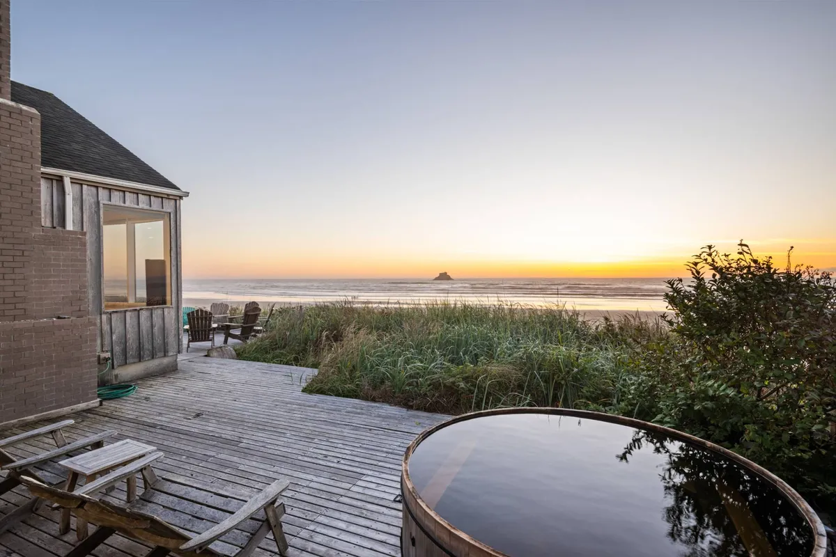 This image features a beautiful wooden deck overlooking the ocean, with a custom hot tub positioned to maximize spectacular sunset views. The scene is tranquil and cozy, capturing both beach grass and the sparkling coastline in the background. The atmosphere is one of relaxation, where guests can soak in the hot tub or gather to watch the sun dip below the horizon.
