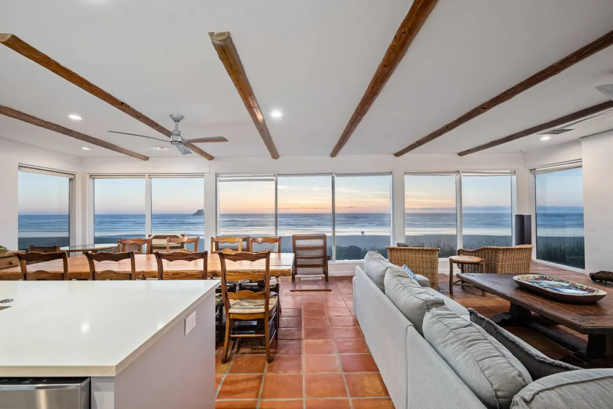 This image provides a wide-angle view of the main gathering zone, featuring a large dining table set beside expansive windows overlooking the Pacific, a welcoming seating area, and a modern kitchen island. The space is bathed in soft natural light and offers a seamless transition between dining, relaxation, and food preparation—all with the breathtaking ocean as a backdrop.