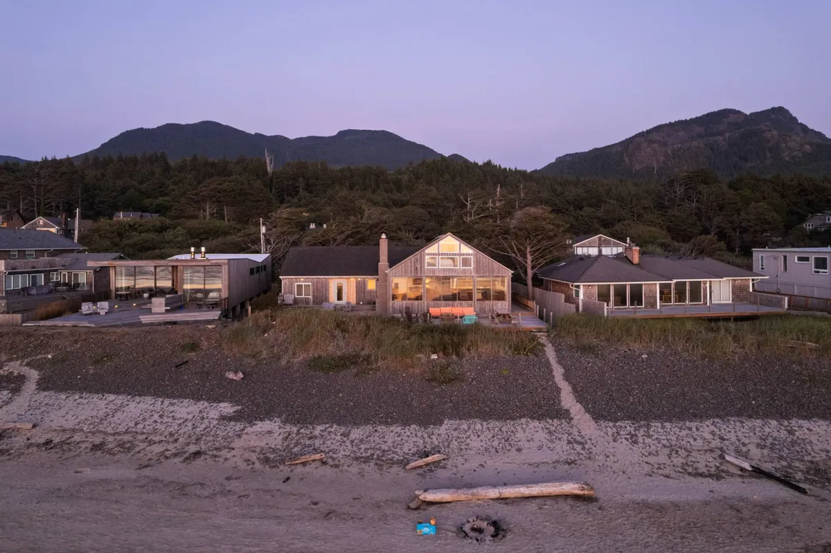 A cozy beachfront house sits against a backdrop of mountains, with a fire pit on the sand and other nearby homes dotted along the shore. The scene captures evening light, inviting gatherings, beach walks, and sunset viewing right at the water's edge.