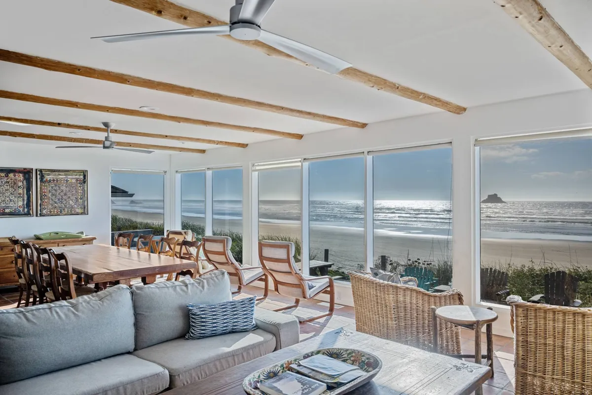 The image showcases a bright, inviting interior living space with expansive windows framing breathtaking ocean views. A spacious dining table sits beside a comfortable seating area, perfect for relaxing together while gazing out at the Pacific. Natural light floods the room, highlighting a seamless blend of indoor comfort and coastal beauty.