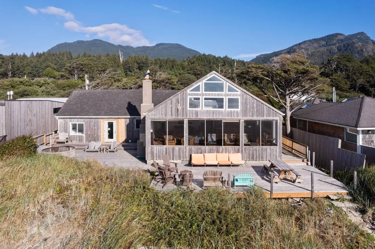 The image captures the large glass front of the house, ample outdoor seating, a visible chimney indicative of a fireplace inside, and lush surroundings. The deck is set up for relaxation with views of the trees and mountains, bridging the indoor and outdoor living experience.