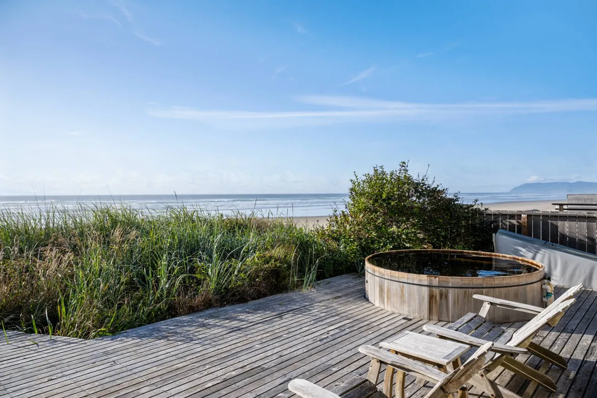 The photo presents a serene hot tub set into a wooden deck with lush greenery around and direct sightlines to the beach and ocean. The setup invites guests to imagine relaxing soaks, sunrise coffee, or stargazing at night, with the sand and surf just steps away.